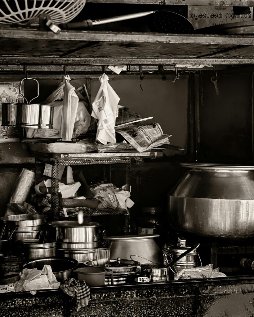 Black and white scene of a traditional kitchen in Kakkanad, India, showcasing cookware and utensils.